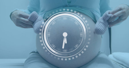 Fast moving clock over midsection of pregnant caucasian woman holding bootees beside belly. Pregnancy, self care, time, development, expectancy, motherhood and lifestyle digitally generated image.の写真素材