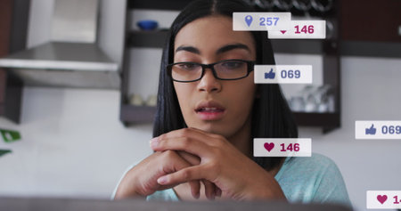 Image of social media icons and numbers over biracial woman using laptop. Global social media and...