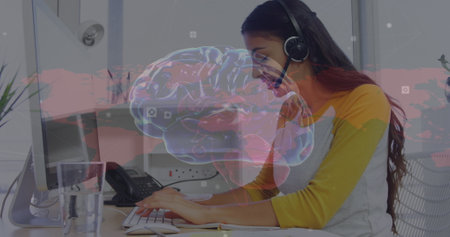 Hispanic woman with headphones types, eyes on screen. She has long brown hair, a yellow top, and is...