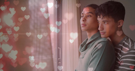 Image of glittering hearts over happy diverse gay male couple looking out of window, embracing. Couple, love, domestic life, lgbtq and lifestyle, digitally generated image.の写真素材