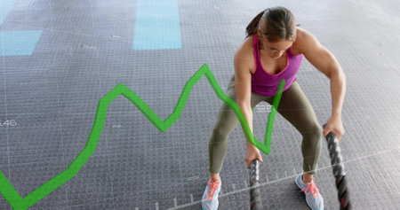 Green line graph image over woman exercising with battle ropes in gym. Fitness, workout, exercise,...