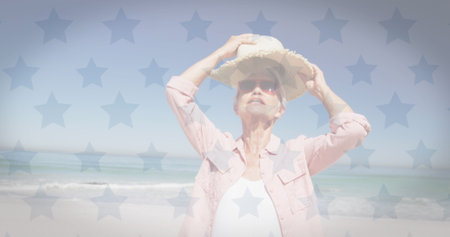 American flag waving over caucasian senior woman wearing a hat at the beach. american independence patriotic holiday conceptの写真素材