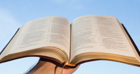 Image of a hand of a person holding open Christian Holy Bible book with blue sky and clouds in the...