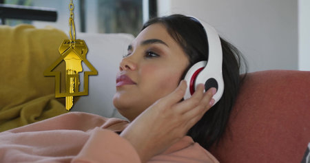 Image of gold house key and key fob over biracial woman using headphones at home. House, home, property, ownership and lifestyle concept digitally generated image.の写真素材