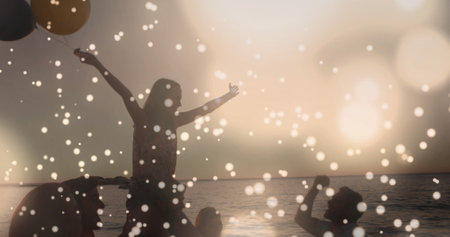 Group of Caucasian friends celebrating at sunset on beach. Holding balloons, throwing confetti, and savoring moment togetherの写真素材