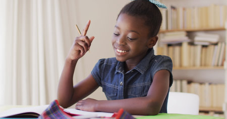 African american girl holding pencil looking out of the window and doing homework while sitting on...