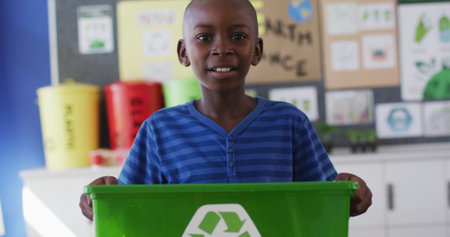 African american schoolboy smiling, holding recycling bin, standing in classroom. children at primary school in summer.の写真素材