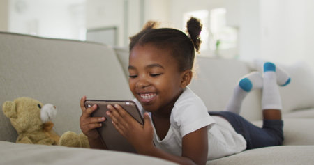 Happy african american girl lying on sofa using digital tablet at home. staying at home in self isolation during quarantine lockdown.の写真素材