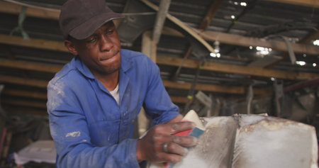 Front view of an African male panel beater in a township workshop, sanding and preparing surface of...