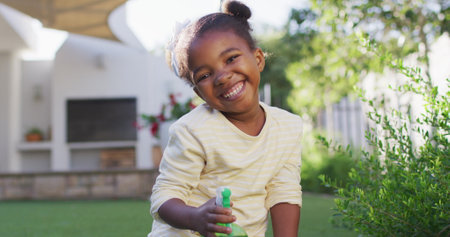 Portrait of happy african amercian girl watering plants holding spray bottle in garden. staying at home in self isolation during quarantine lockdown.の写真素材