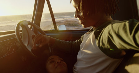 Front view of African american woman lying on mans lap in a pickup truck at beach. They are talking...