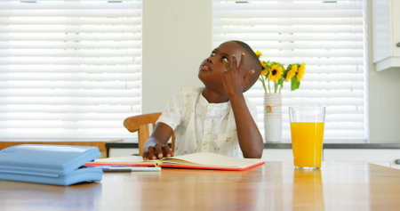 Front view of little black boy doing homework at dining table in a comfortable home. He is counting with his fingers and looking up 4kの写真素材
