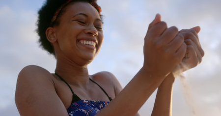 Low angle view of African american woman playing with sand on the beach. She is smiling and having fun 4kの写真素材