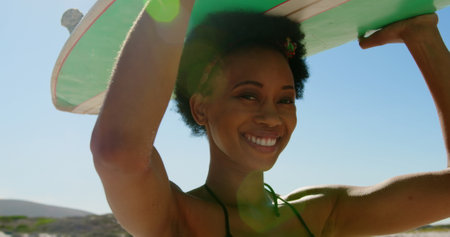 Close-up of young African American female surfer smiling and looking at the camera while carrying surfboard on her head at beach 4kの写真素材