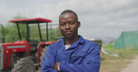 Portrait of a young African American male farmer on an organic farm, standing in front of a tractor with arms crossedの写真素材