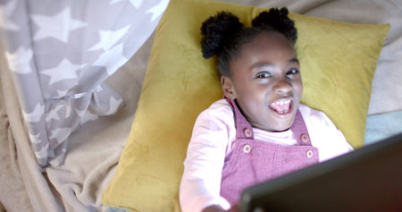 Happy african american girl lying in tent and using tablet at home, slow motion. Lifestyle, childhood, free time, communication and domestic life.の写真素材