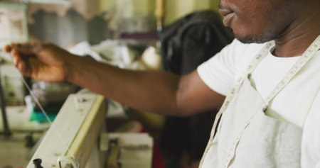 Side view of an African male tailor in a township workshop, sitting by a sewing machine and cutting a thread, slow motionの写真素材