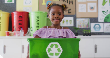 Happy african american schoolgirl standing in classroom, smiling, holding recycling bin. children in primary school.の写真素材