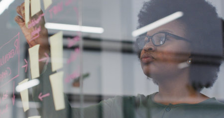 Smiling african american businesswoman brainstorming using memo notes on glass wall in office. work at an independent creative business.の写真素材