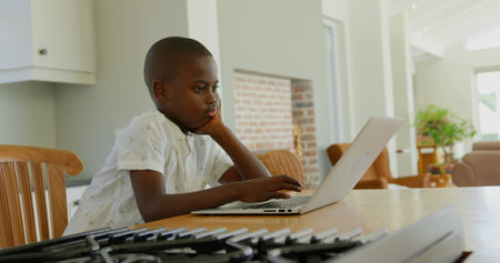 Side view of little black boy using laptop at dining table in a comfortable home. He is leaning his...