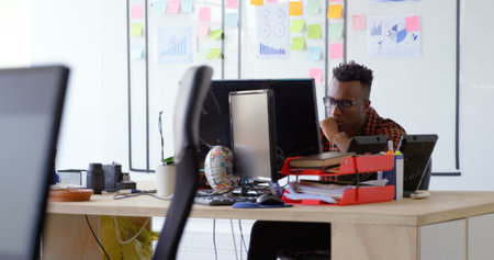 Front view of thoughtful young black businessman working and sitting at desk in a modern office. He is looking at computer screenの写真素材