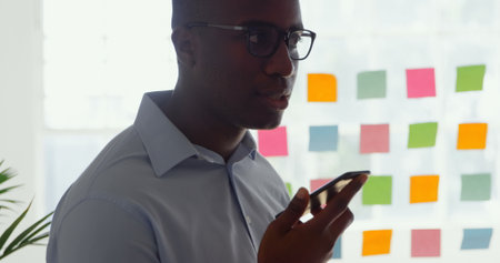 Side view of young black businessman talking on mobile phone in a modern office. Sticky notes on a...