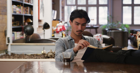 Front view of handsome biracial man with short dark hair enjoying time at home, sitting with his feet up on a table in his sitting room, reading a book, cup of coffee next to him.の写真素材