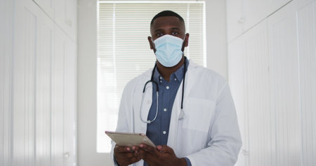 African american doctor wearing face mask using digital tablet at home. distant communication and telemedicine consultation during covid-19 pandemic concept.の写真素材