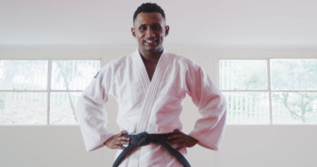 Portrait of a happy confident teenage biracial male judoka wearing white judogi, standing in the gym...