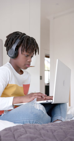 Vertical image of happy african american boy sitting on bed using laptop at home. Childhood, communication and domestic life, unaltered.の写真素材