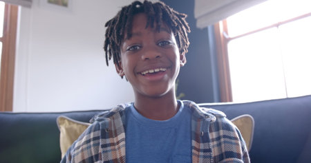African american boy sitting on sofa having image call at home, slow motion. Childhood, communication and domestic life, unaltered.の写真素材