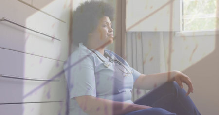 Image of rain on window over unhappy african american female doctor sitting in corridor. Sadness, work, stress, depression, healthcare services and mental health, digitally generated image.の写真素材