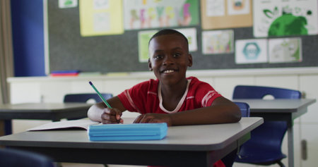 Portrait of happy african american schoolboy sitting at classroom, making notes, looking at camera....