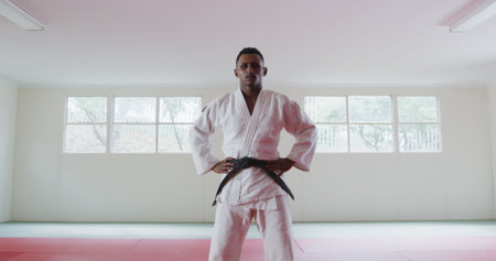 Portrait of a confident teenage biracial male judoka wearing white judogi, standing in the gym with his hands on hips and looking straight into a cameraの写真素材