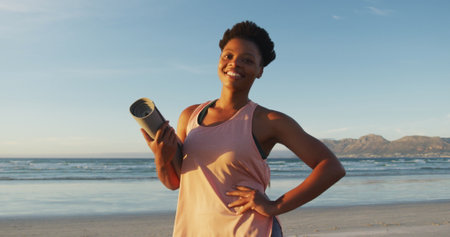 Portrait of african american woman holding yoga mat at the beach smiling. healthy active lifestyle, outdoor fitness and wellbeing.の写真素材