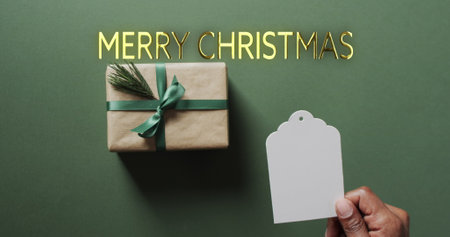 Merry christmas text in gold with gift and african amrican male hand holding blank tag on green. Christmas, tradition, greetings and celebration digitally generated image.の写真素材