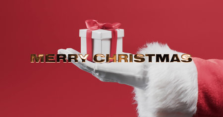 Merry christmas text in gold over hand od santa claus holding present on red background. Christmas, tradition, greetings and celebration digitally generated image.の写真素材