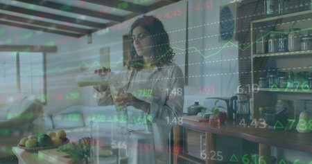 Image of financial data processing over biracial woman making healthy smoothie in kitchen. Global...
