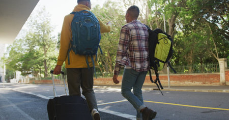 Back view of two happy biracial male friends walking and talking down the street. backpacking...