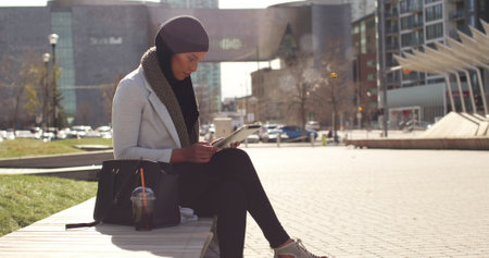Urban professional woman using tablet and enjoying iced coffee on city bench. Business, technology,...