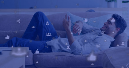Relaxing on couch, young man browsing smartphone with digital icons overlay. Technology, relaxation, connectivity, social media, communication, leisureの写真素材