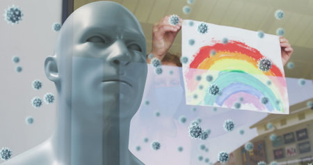 Image of covid 19 cells over model head with person in face mask holding rainbow drawing. healthcare and protection during coronavirus covid 19 pandemic, digitally generated image.の写真素材
