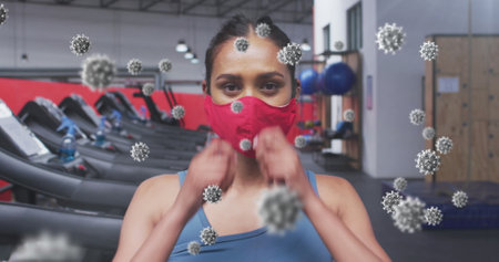 Multiple covid-19 cells floating against portrait of woman wearing face mask at the gym. coronavirus covid-19 pandemic and fitness conceptの写真素材