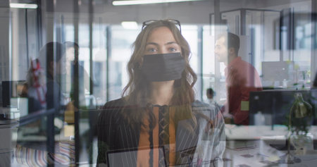 Composite of caucasian woman wearing face mask in office, and colleagues talking. business and workplace hygiene during covid pandemic concept digitally generated image.の写真素材