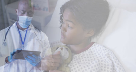 Composite of male doctor in face mask using tablet, and girl patient lying in hospital bed. medical services, technology and healthcare during covid pandemic concept digitally generated image.の写真素材