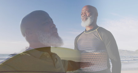 Composite of african american senior man stretching arms, and carrying surfboard on sunny beach. healthy retirement, fitness and wellbeing concept digitally generated image.の写真素材