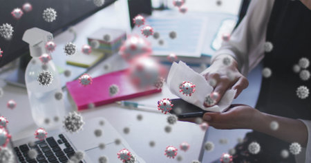 Image of covid 19 cells floating over woman wiping smartphone in office. workplace and business during covid 19 pandemic concept digitally generated image.の写真素材