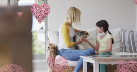 Image of hearts over caucasian woman and her daughter wearing face masks playing with pet dog. family time at home during covid 19 pandemic digitally generated image.の写真素材