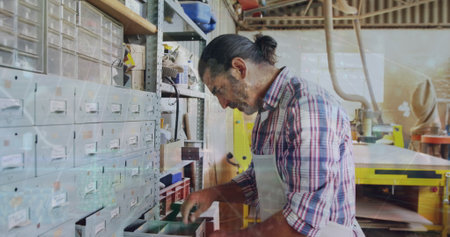 Craftsman wearing apron sorting components from storage drawers in workshop, with yellow machine....