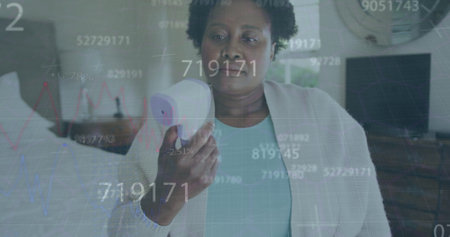 Multiple changing numbers over african american senior woman measuring her temperature at home. Covid-19 pandemic conceptの写真素材